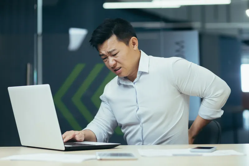 Office worker experiencing lower back pain while using a laptop at a desk after prolonged sitting.