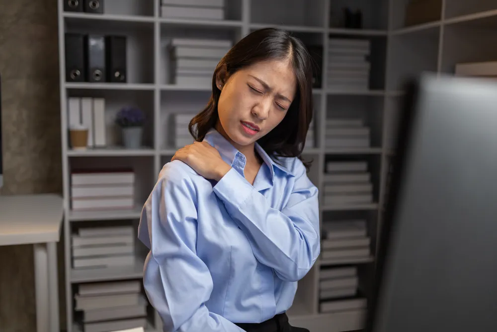 Office worker holding her shoulder while sitting at a desk, depicting neck and shoulder pain associated with prolonged computer use and office syndrome.