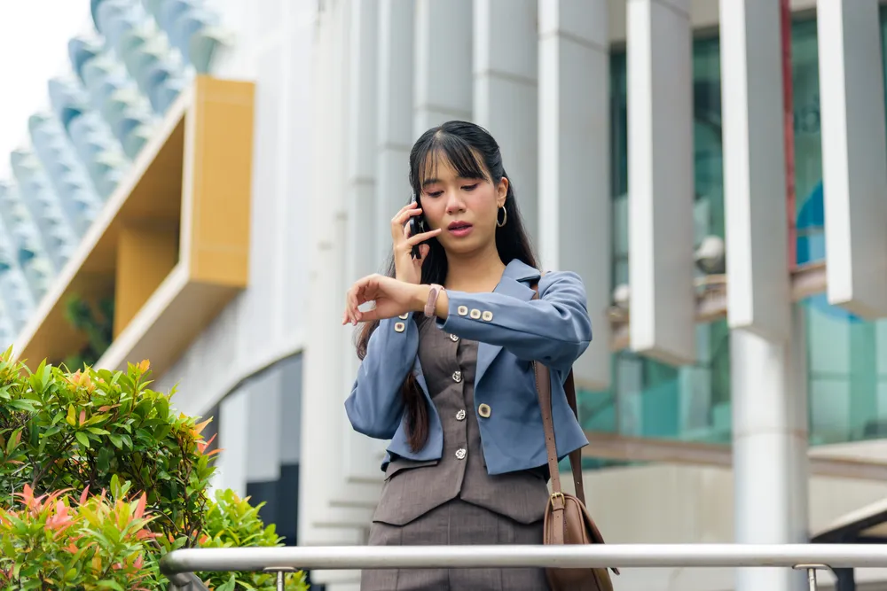Businesswoman checking her watch while talking on her phone outside an office building in Singapore, appearing tired after a busy workday.