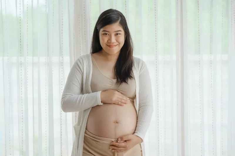 Pregnant Asian woman smiling while gently holding her baby bump indoors
