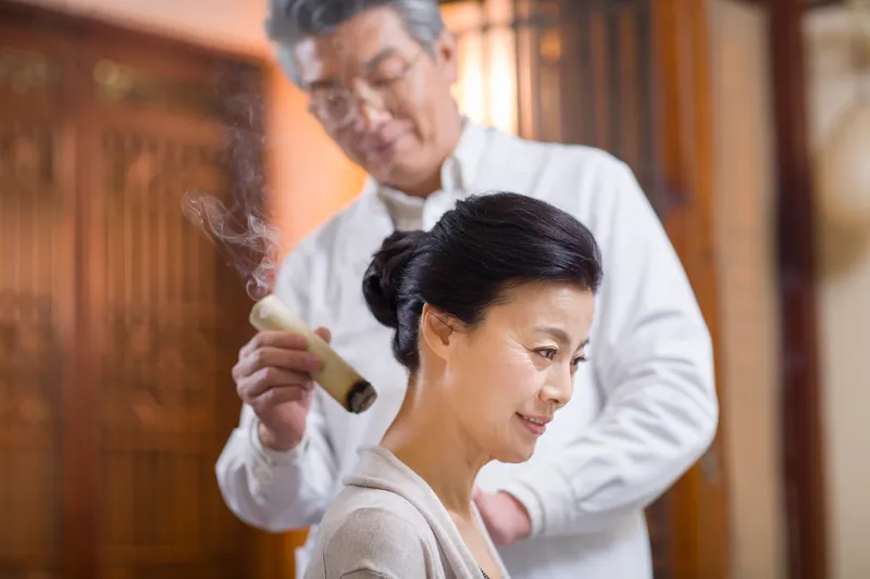a tcm practitioner giving a woman a moxibustion therapy for her neck pain